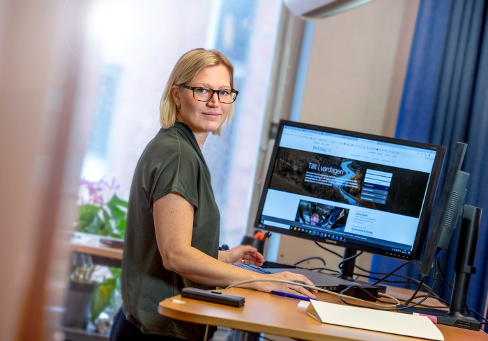 Martina står vid sin dator och ler mot kameran
